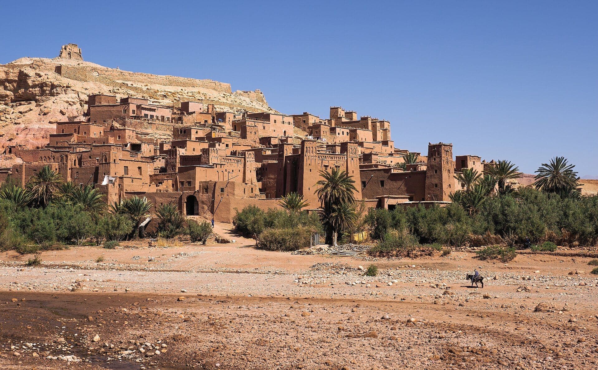 The mud-brick ksar of Aït Benhaddou against a hill, with the riverbed and palm trees in the foreground