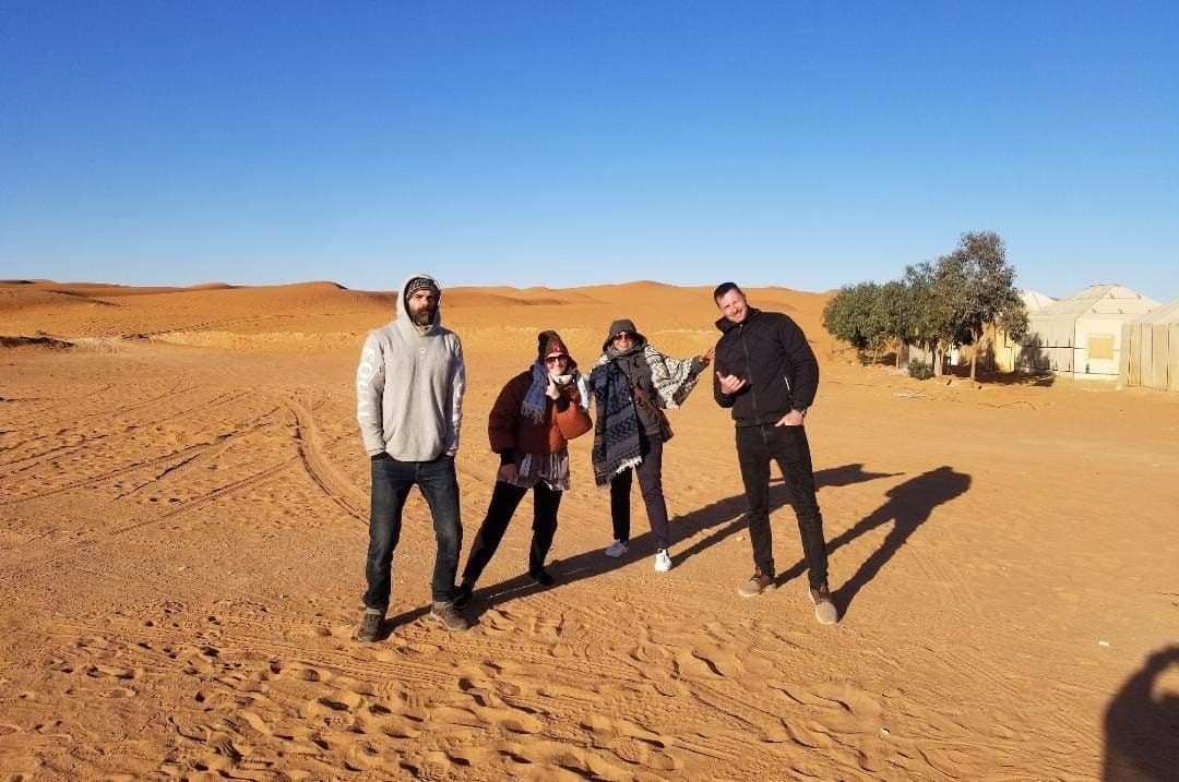 Four travelers pose in the sand dunes near a desert camp