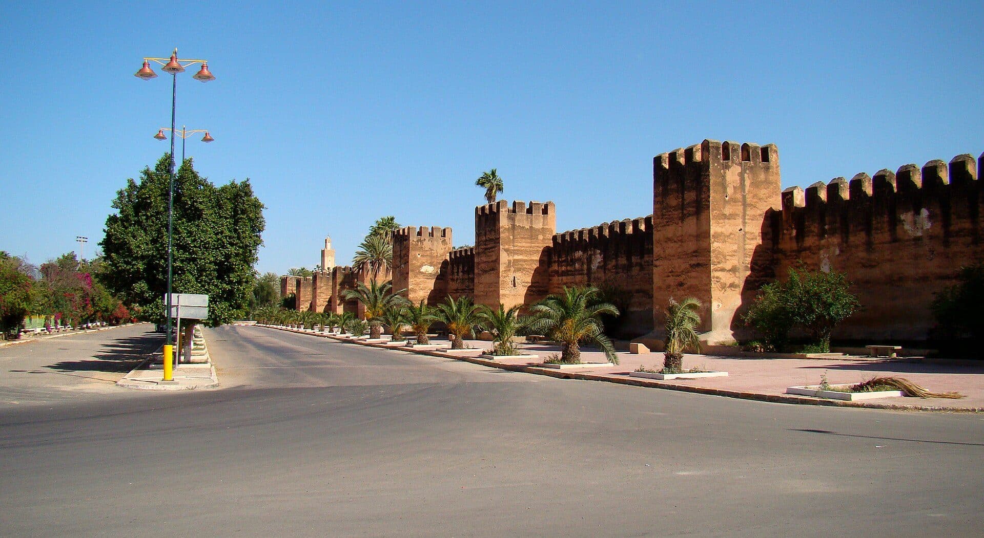 De lemen stadsmuur van Taroudant met kantelen en vierkante torens, met palmen en een brede weg ervoor