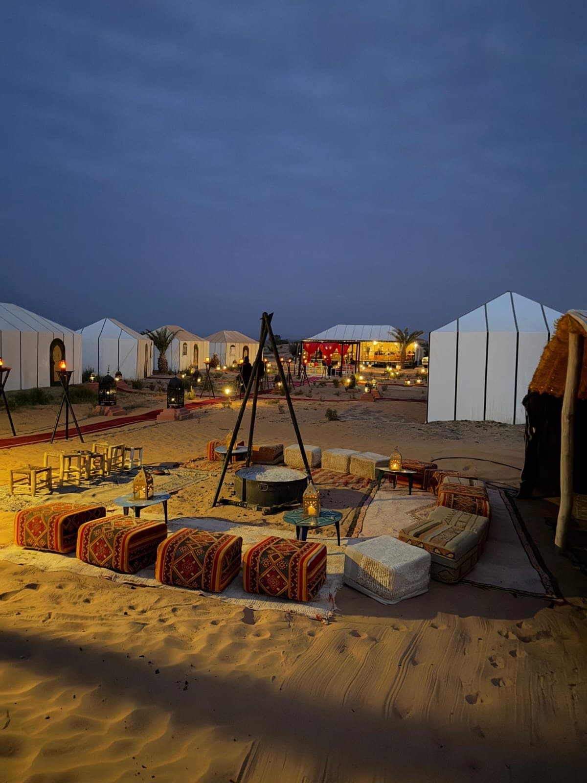 Witte tenten en zitkussens rond een vuurplaats in een woestijnkamp bij Merzouga, in de schemering